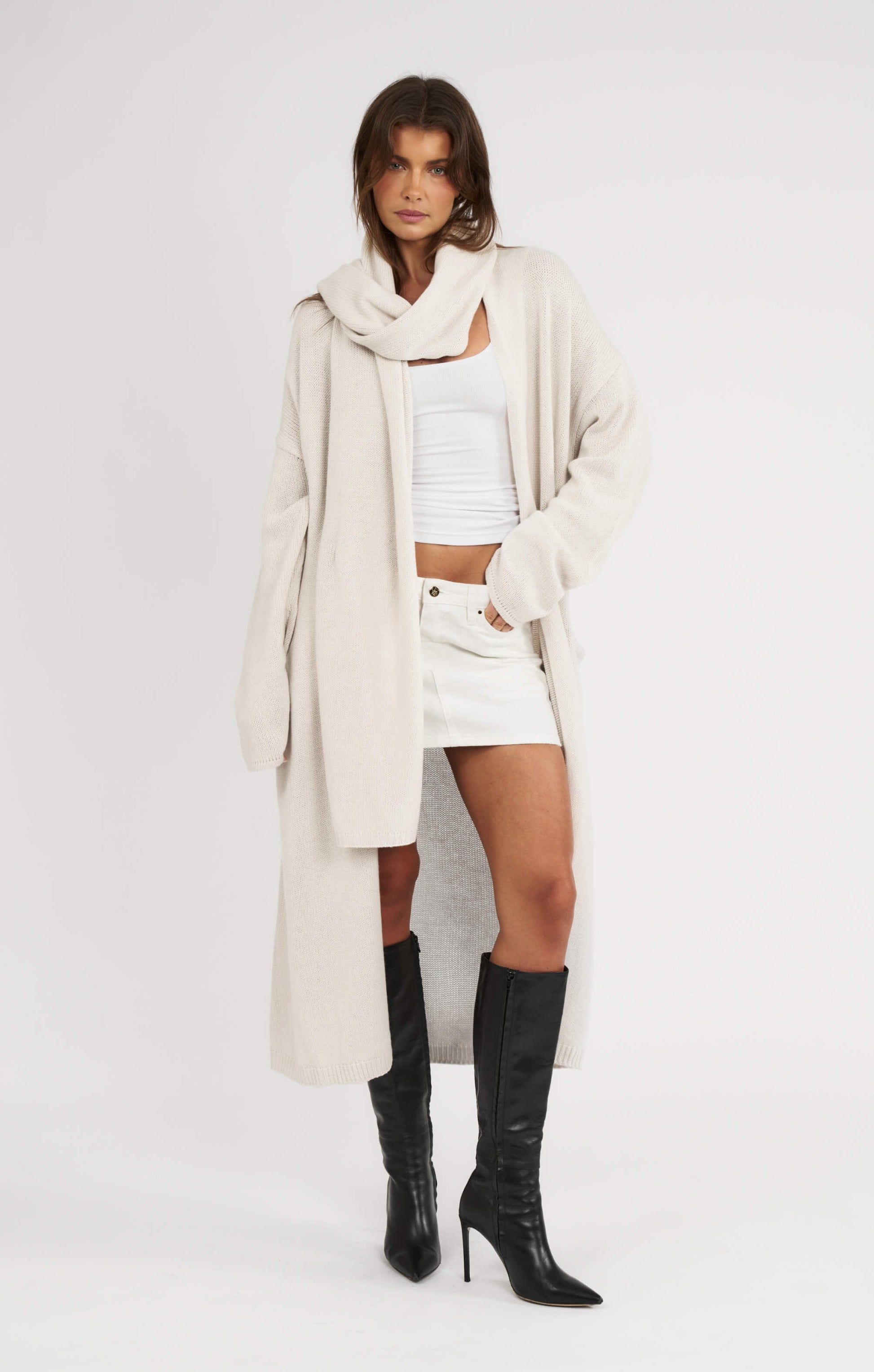 Woman wearing a long beige coat, white top, and black knee-high boots on a white background