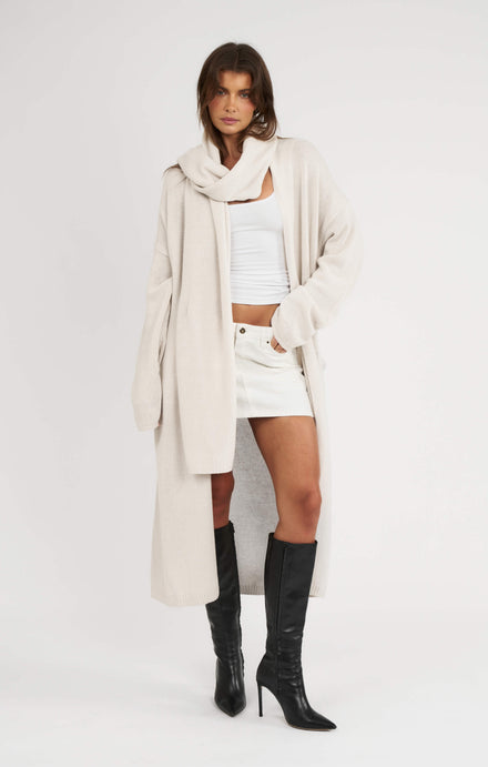 Woman wearing a long beige coat, white top, and black knee-high boots on a white background